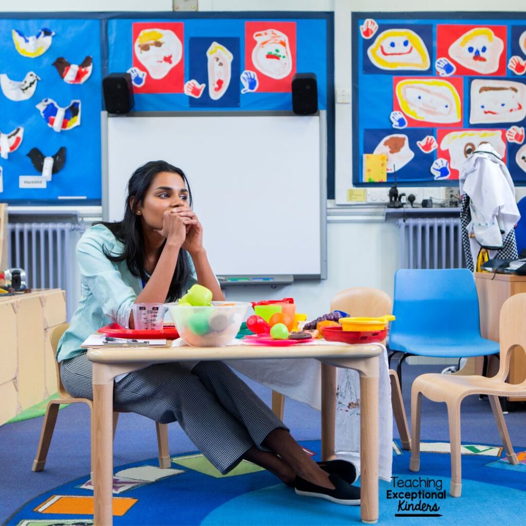 A teacher sits at a cluttered classroom table, looking overwhelmed.